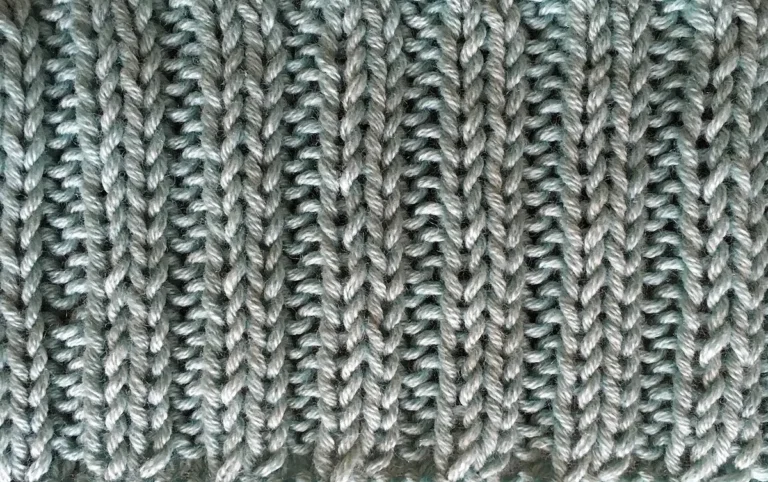 Close view of a hand-knitted grey 2x2 rib swatch showing alternating raised and recessed vertical columns